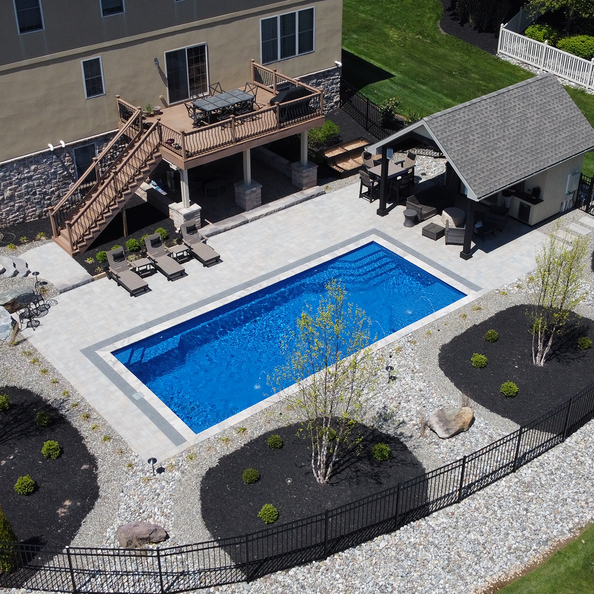 An aerial view of a backyard with a swimming pool, poolside lounge chairs, a deck with outdoor furniture, a small building, and landscaped areas with trees and rocks.