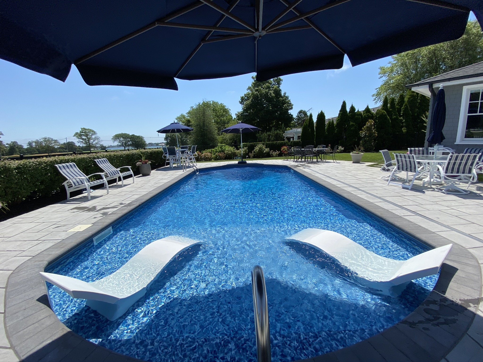 Backyard with a swimming pool, lounge chairs, patio tables, umbrellas, and trees under a clear blue sky.