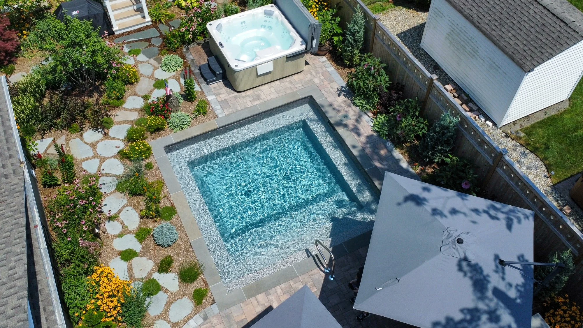 A backyard view from above features a small square swimming pool, a hot tub, and a garden with flowers and stepping stones.