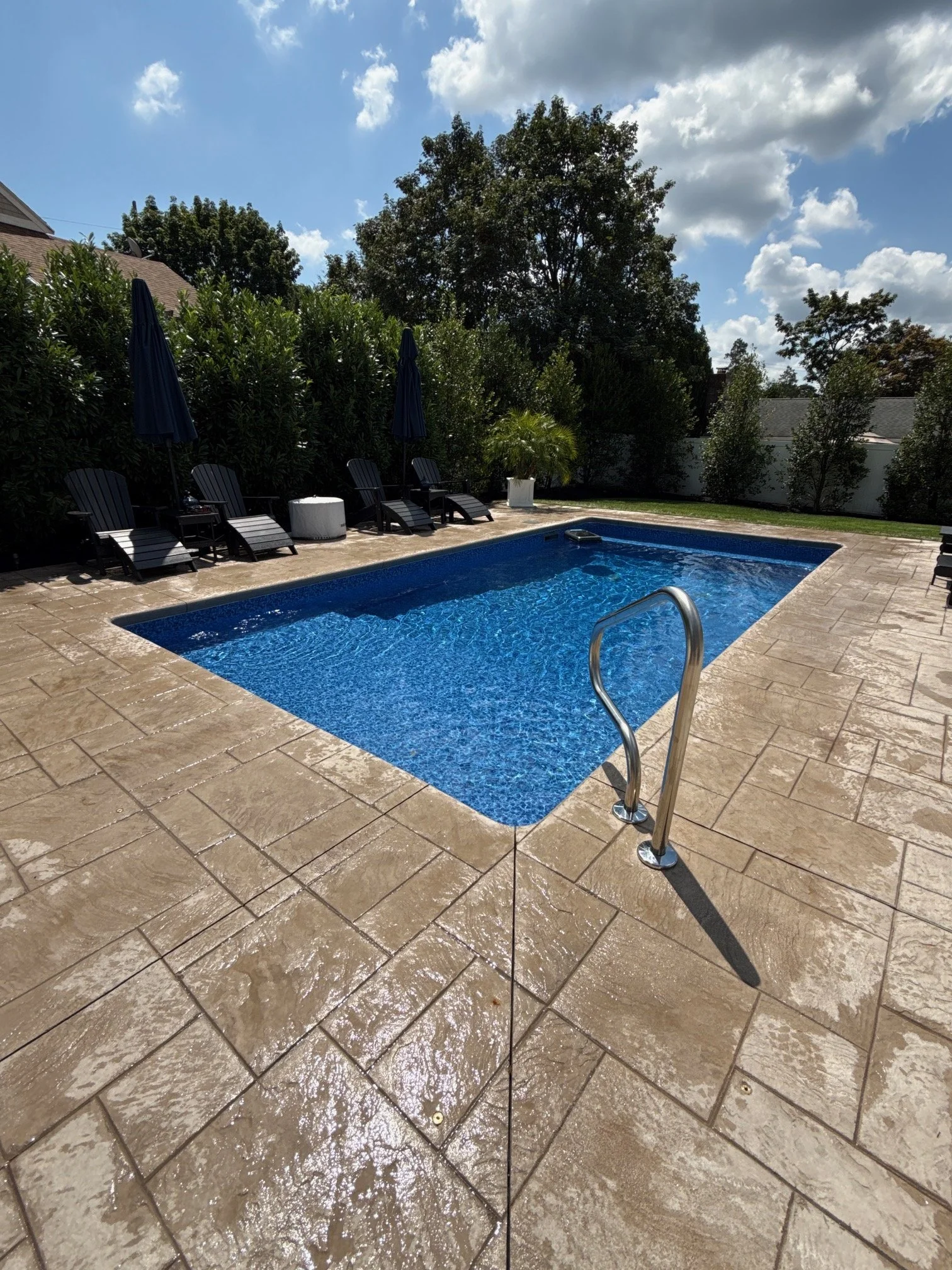 A backyard with a small swimming pool, surrounded by wet stone tiles and several lounge chairs with umbrellas, lush green bushes, and trees under a partly cloudy sky.