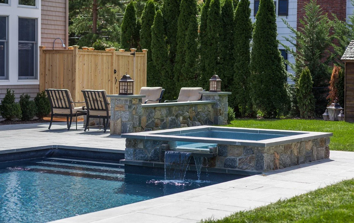 Modern backyard with swimming pool, hot tub, patio furniture, stone accents, and lush green trees and bushes in the background.