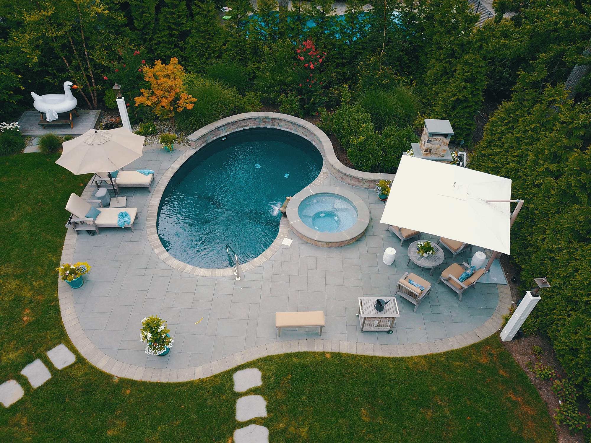 A backyard with a kidney-shaped pool, hot tub, and outdoor seating area shaded by large white umbrellas. The yard is surrounded by dense green trees and landscaping, with a small wooden deck featuring a large inflatable swan and a garden light.