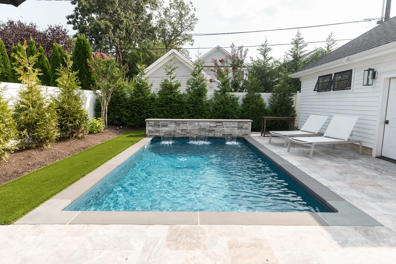 Backyard with a rectangular swimming pool, two white lounge chairs, a stone water feature with three water spouts, green shrubs and trees, and a white house with awning windows.