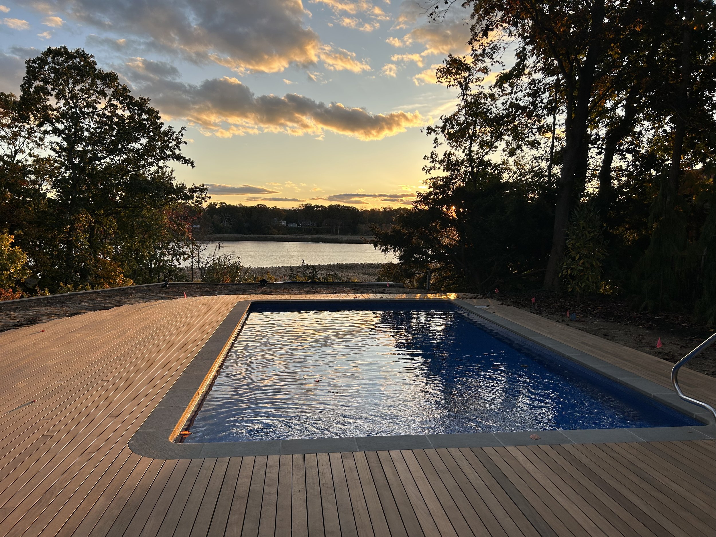 An outdoor swimming pool with a wooden deck, surrounded by trees and overlooking a body of water in the distance during sunset.