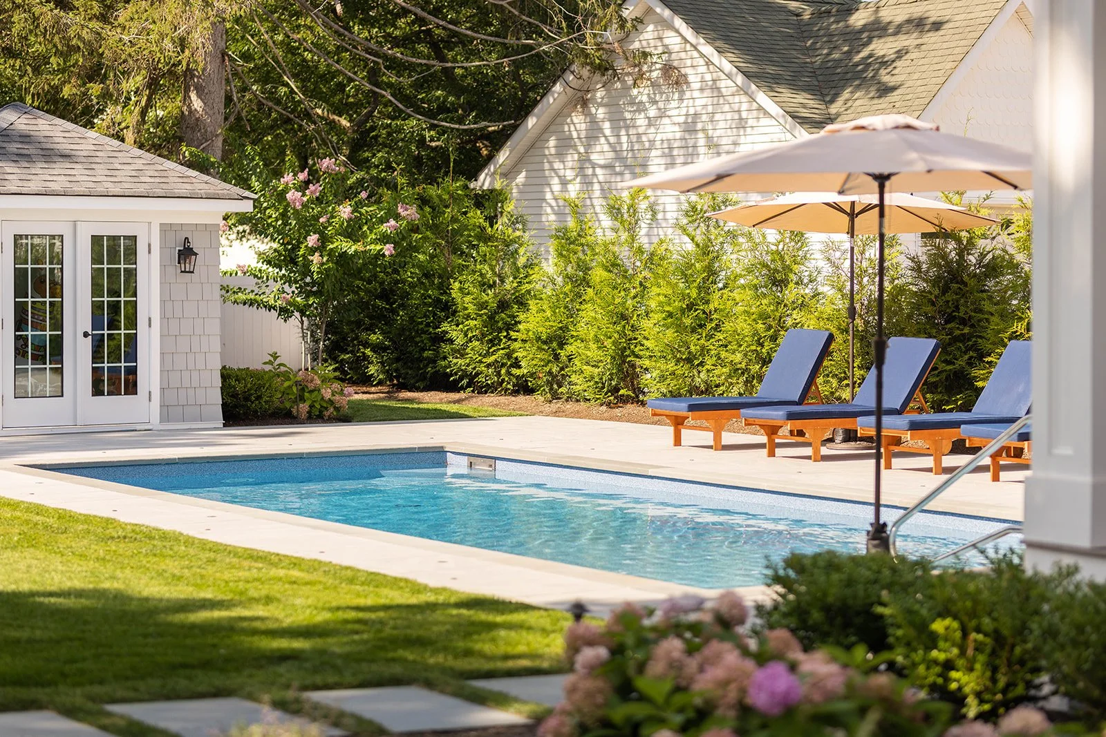 A backyard with a swimming pool, two blue lounge chairs with a small table, and a large umbrella for shade. There are bushes and trees surrounding the area, with a small shed and a house in the background.