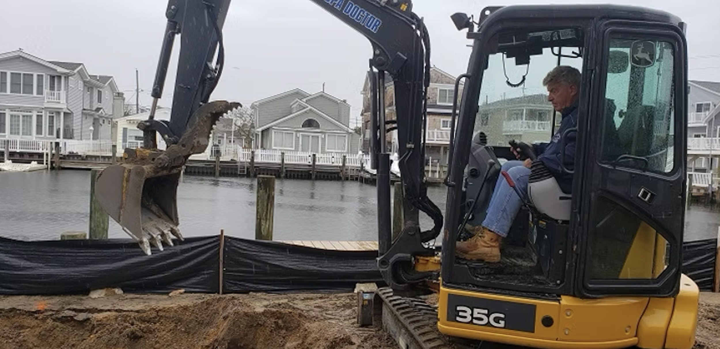 A man operating a small yellow and black excavator near a waterfront with houses on the other side of the water, overcast sky.