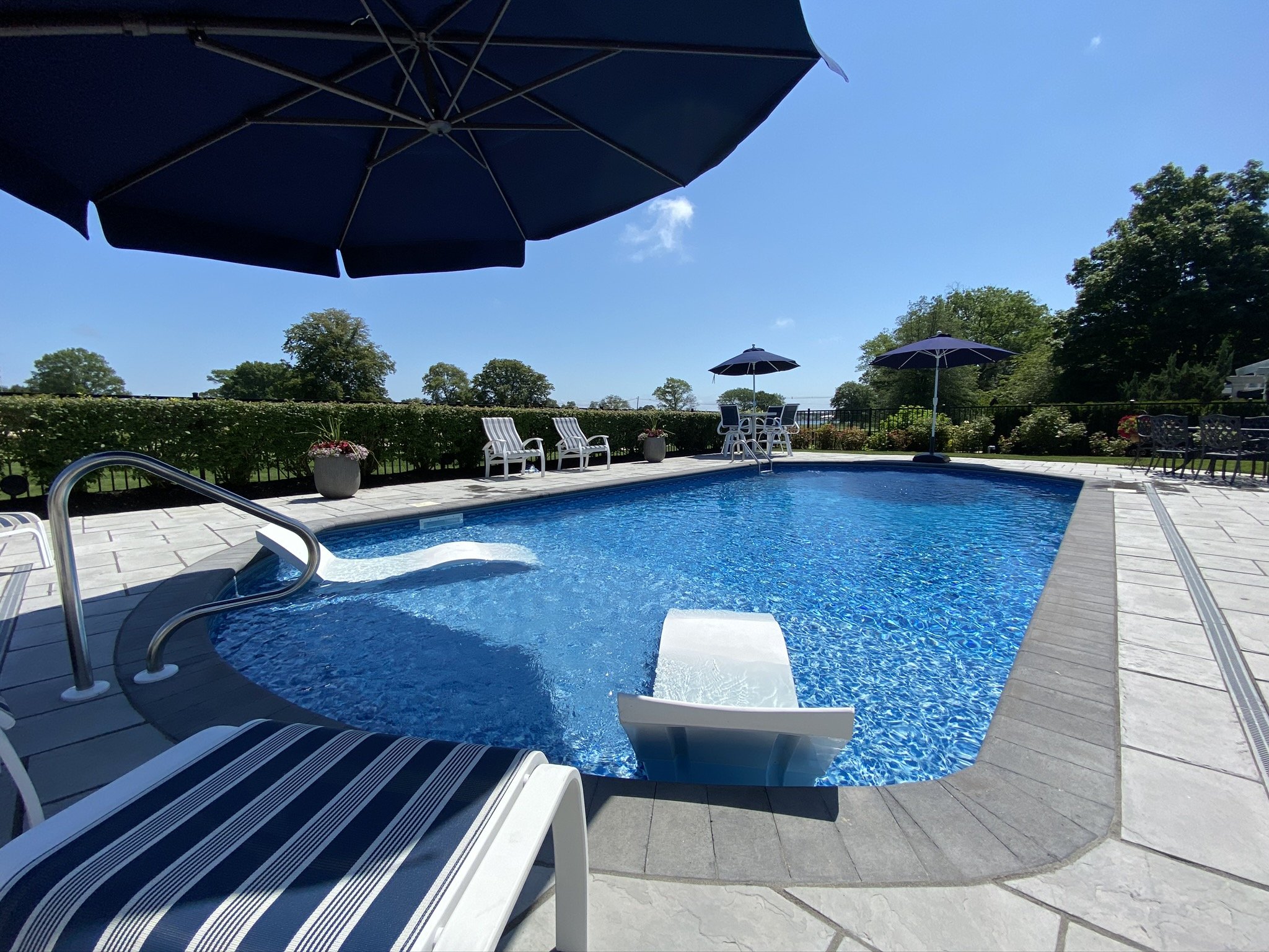 Swimming pool with lounge chairs and umbrellas on a sunny day, surrounded by trees and clear blue sky.