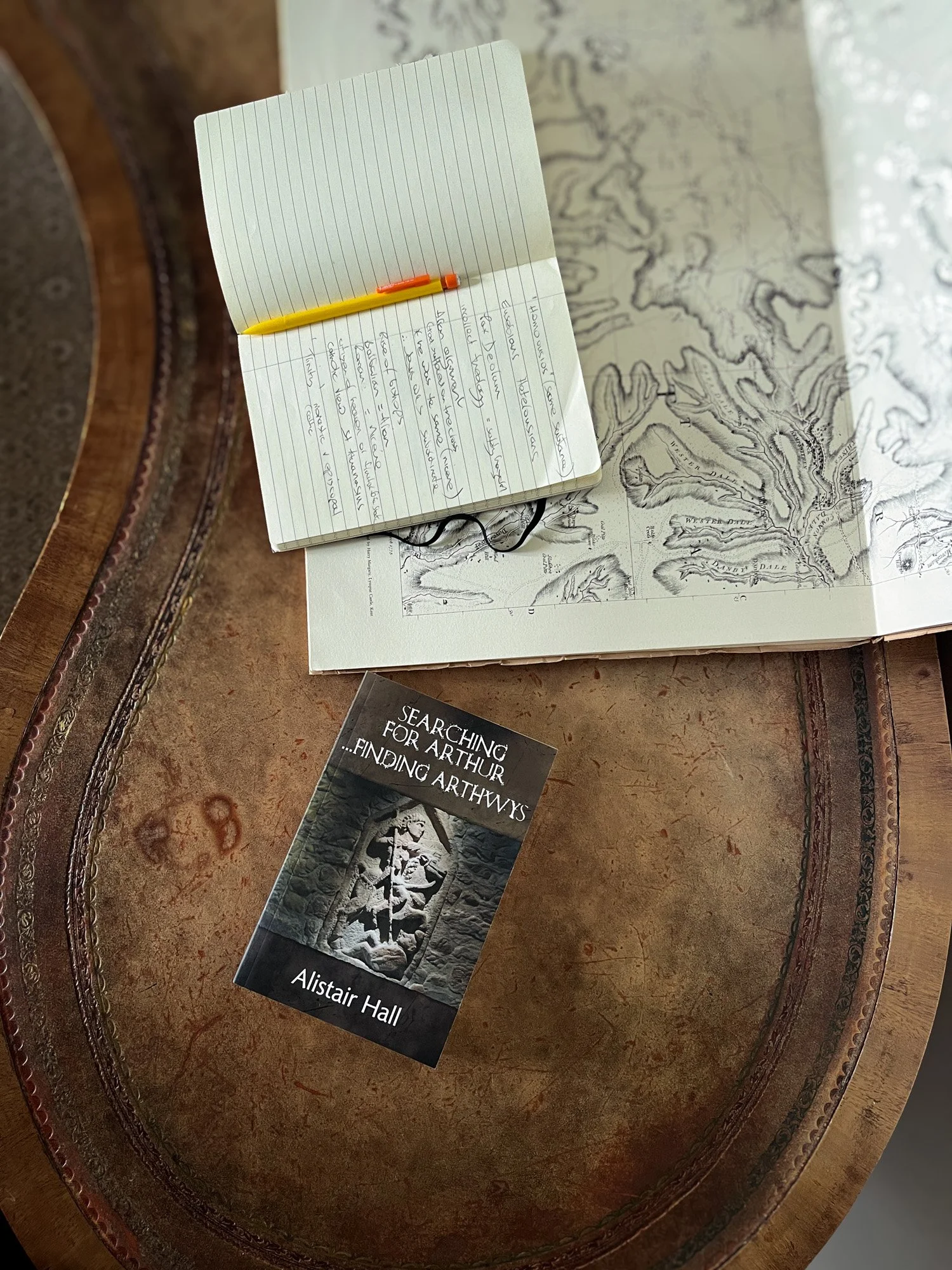 The book Searching for Arthur...  finding Arthwys is seen from above lying on a brown desk next to a large hand drawn map with an open notebook on it. There is a pen in the crease of the notebook.