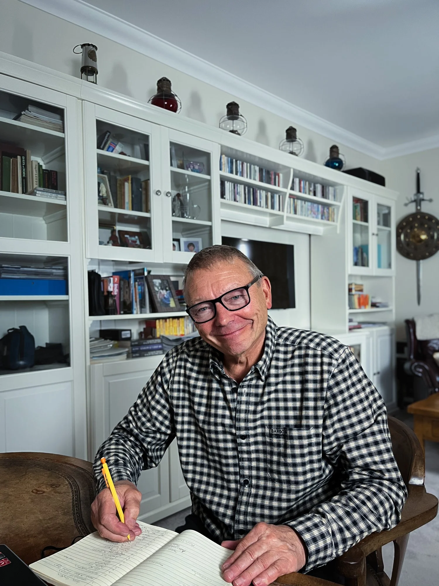 Author Alistair Hall writing in a notebook at his desk. In the background are shelves with books.