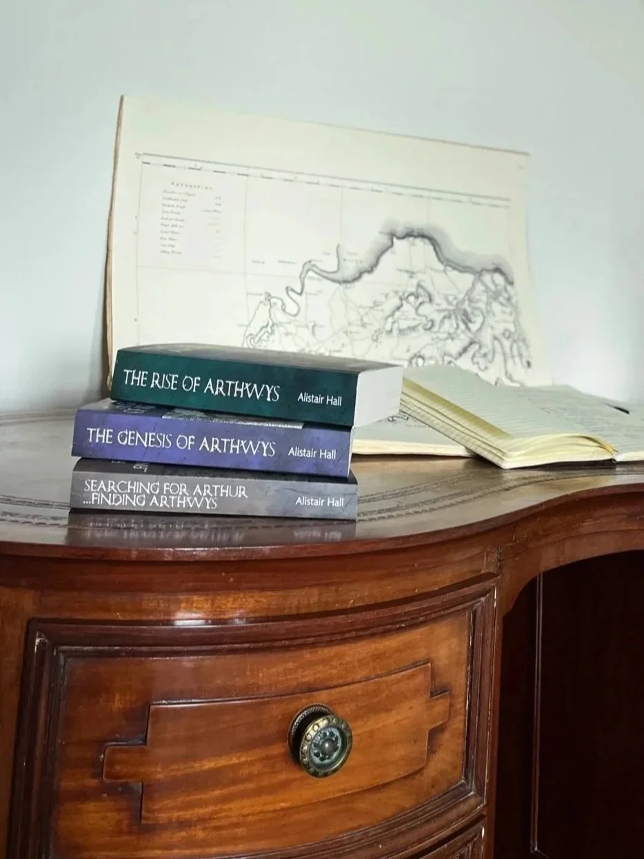 A wooden desk with three books written by author Alistair Hall in a pile. Behind the books leaning against the wall is a large old map with a notebook open in front of it.