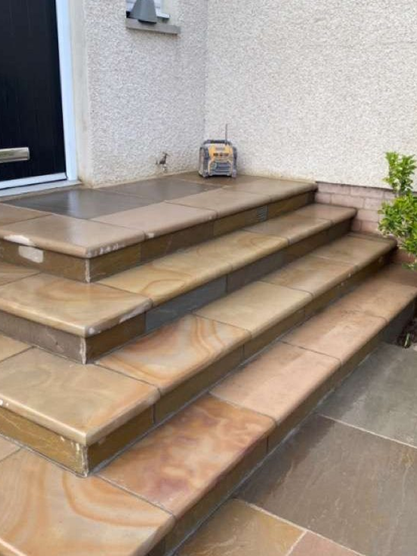 Four-tiered outdoor staircase with tan and brown tiles, leading up to a black door, with a small potted plant on the right
