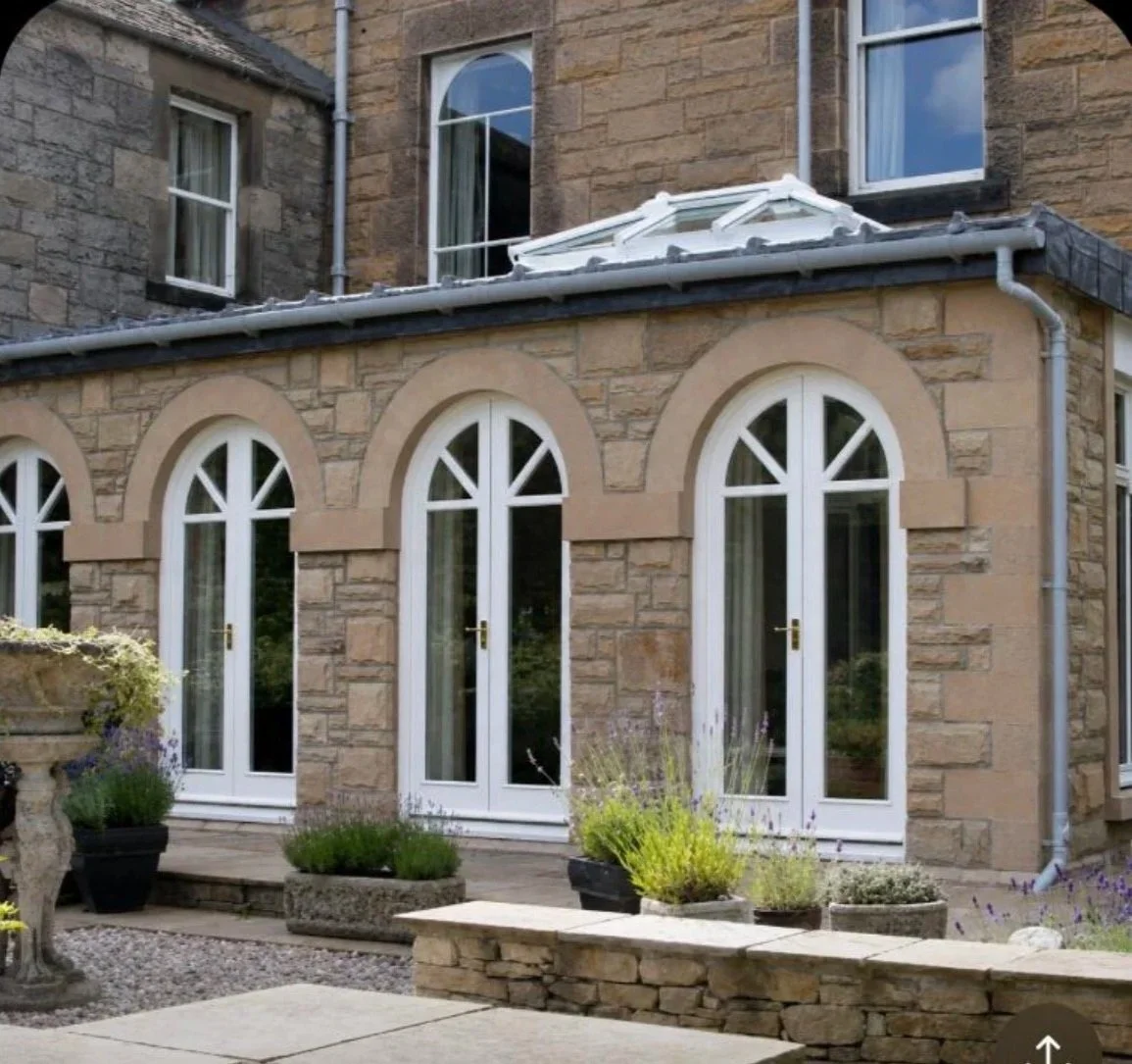 Back of a stone house with arched windows and a small extension with glass doors, potted plants, and a stone wall.
