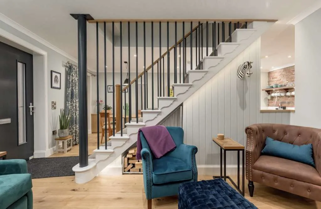 Living room with a staircase, blue and brown armchairs, a small wooden side table, and a fireplace in the background.