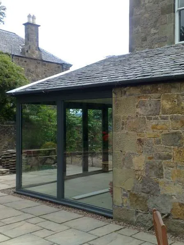 A glass-enclosed extension with a sloped roof attached to a stone building in a garden setting.