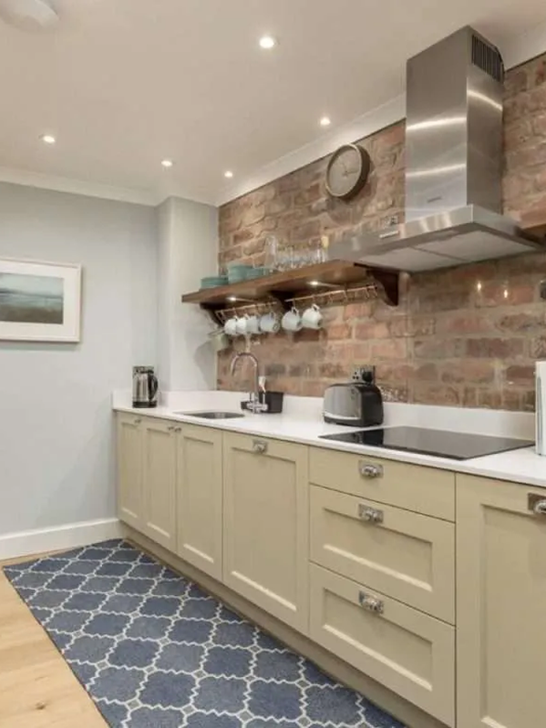 Kitchen with beige cabinets, white countertop, brick wall behind, open wooden shelf with cups and glasses, stainless steel range hood, clock, coffee maker, toaster, electric stove, patterned blue rug, and framed picture on gray wall.