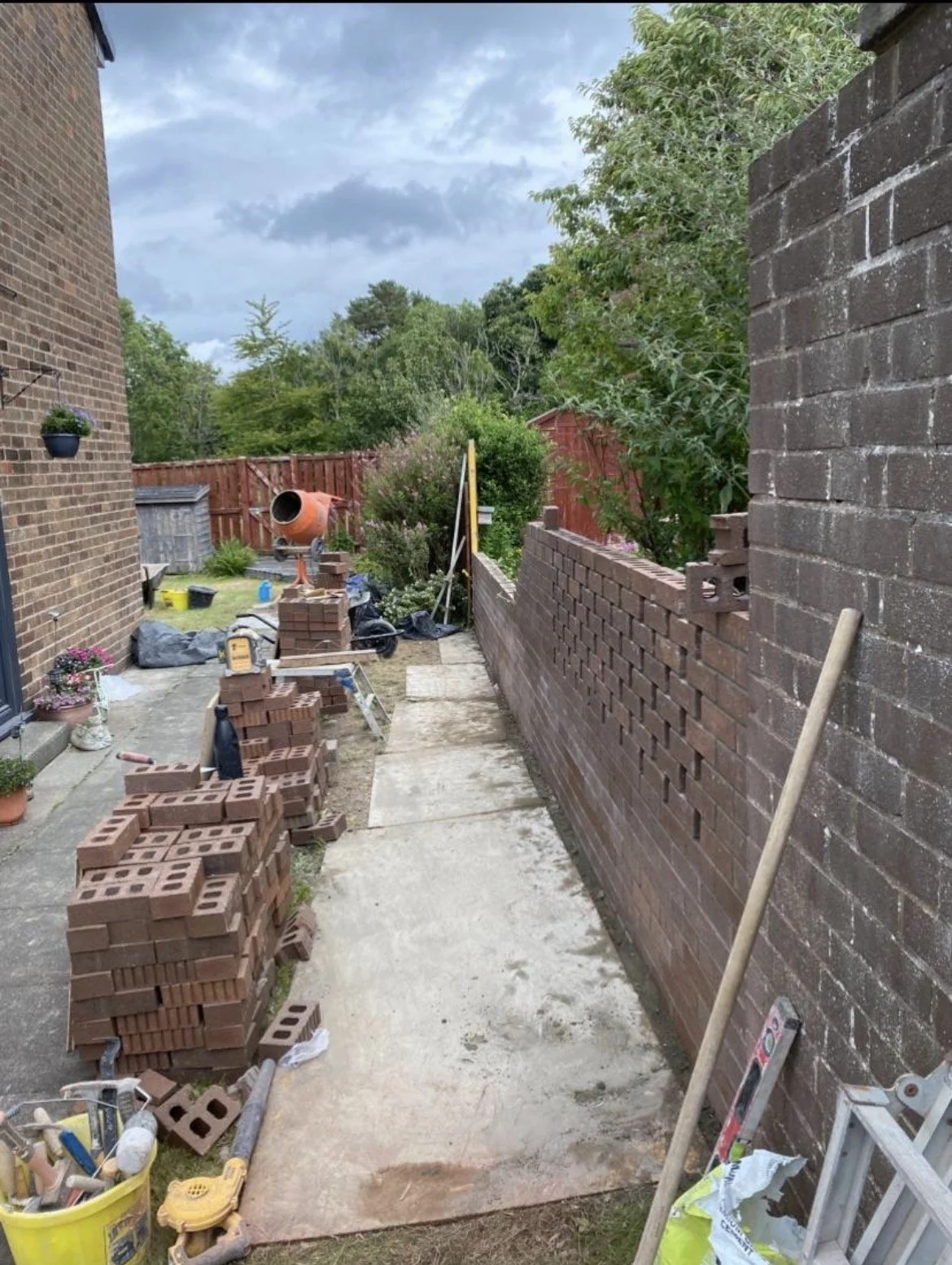 Backyard construction site with brick wall being built, construction tools, stacked bricks, and a concrete mixer in the background, surrounded by trees and cloudy sky.