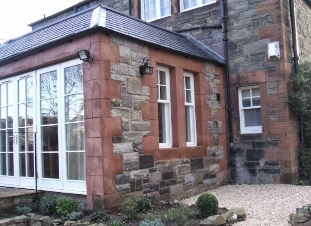 A stone and brick house with a conservatory on the left side, three windows with white frames on the main wall, and a gravel and stone garden area in front.