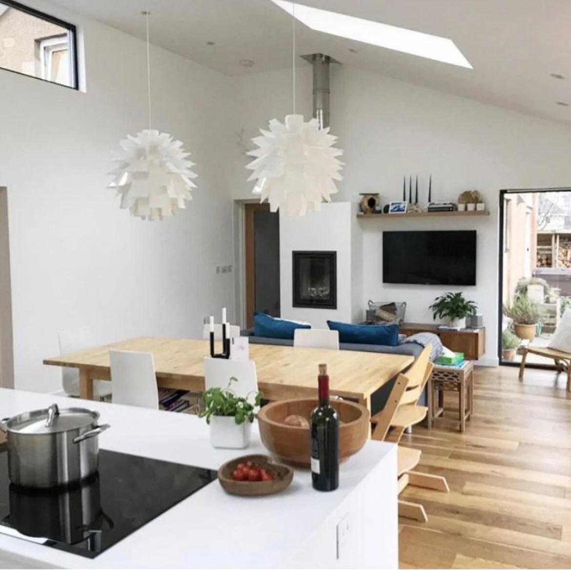 Open-concept living and dining area with a white kitchen island, wooden dining table, and modern white and wooden chairs. Hanging pendant lights over the dining area, a wall-mounted TV, a fireplace, and a cozy seating area with blue cushions. Large windows and a patio door open to an outdoor space with potted plants.
