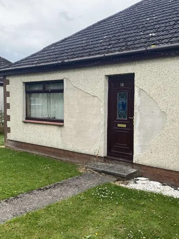 Front of a house with a damaged wall near the front door, showing peeling plaster and cracked paint, with a window to the left and a dark-colored door with a glass panel.