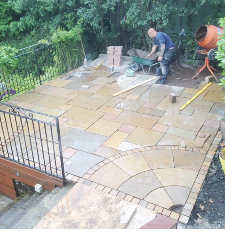 A person laying stone pavers on a patio outdoors, with construction tools like a wheelbarrow, level, and cement mixer nearby.