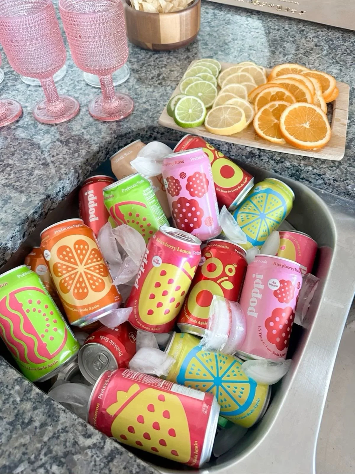 A kitchen countertop with four pink textured glasses, a wooden bowl, a cutting board with lemon, lime, and orange slices, and a large tray filled with colorful cans of soda and some ice cubes.