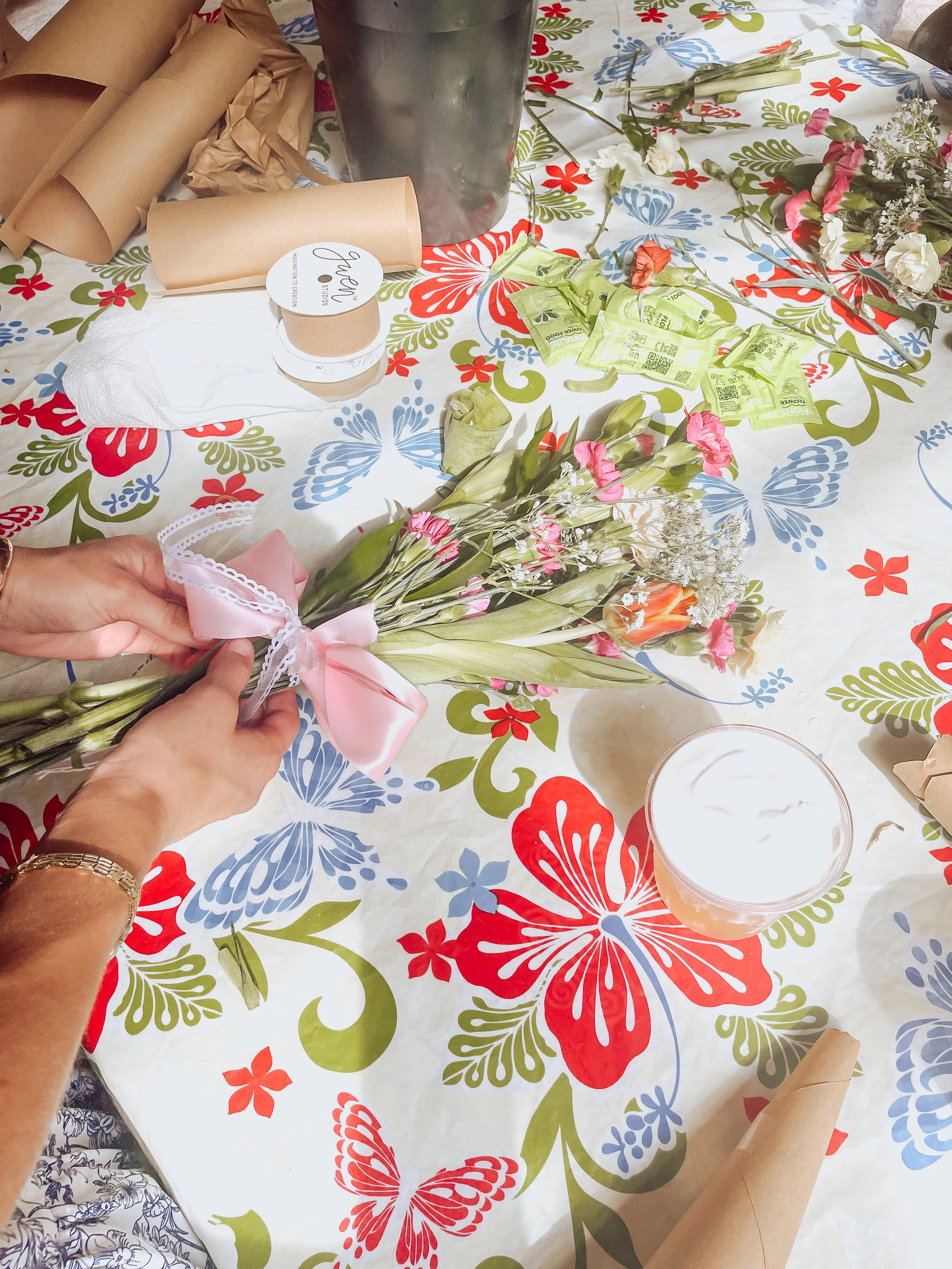 A table covered with a colorful floral tablecloth, with flowers, green packets, brown paper wrapping, a black cup, a white napkin, and a person's hands holding a small bouquet of pink, white, and peach flowers with a pink ribbon.