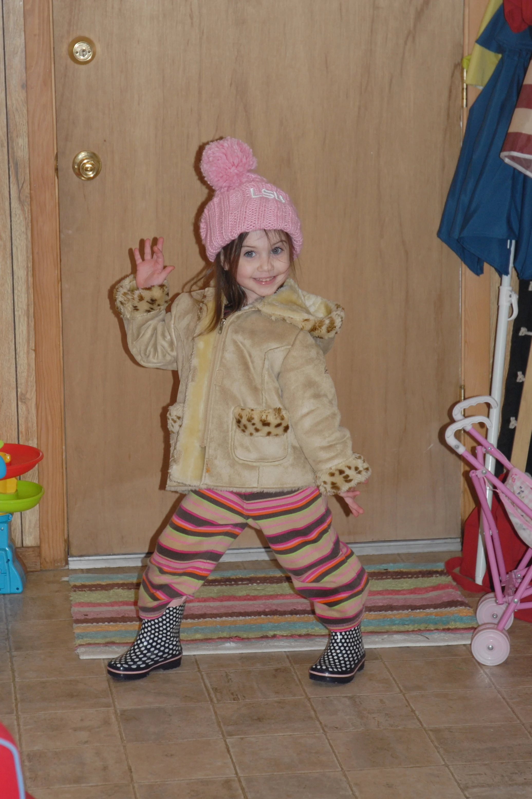 Cute young girl in a colorful striped pajama pants and black-and-white polka dot rain boots posing playfully inside a house, wearing a pink knit beanie with a pom-pom, and a beige faux fur coat with leopard print accents, standing in front of a wooden door with a blue coat hanging on the side.