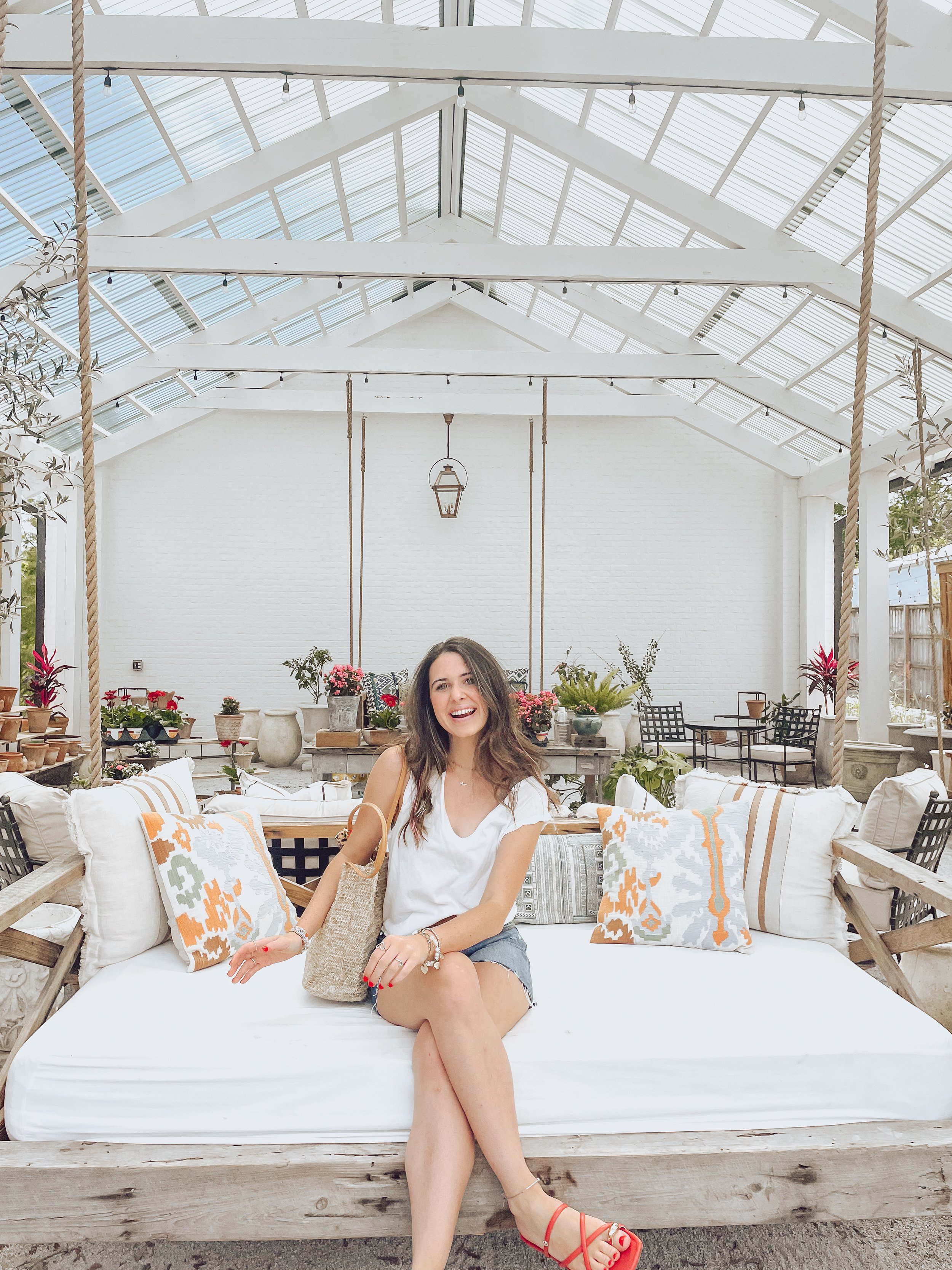 A young woman with long dark hair, sitting on a white outdoor sofa with patterned pillows, smiling and holding a beige tote bag. The room has a glass roof, plants, hanging swing ropes, and a white brick wall in the background.