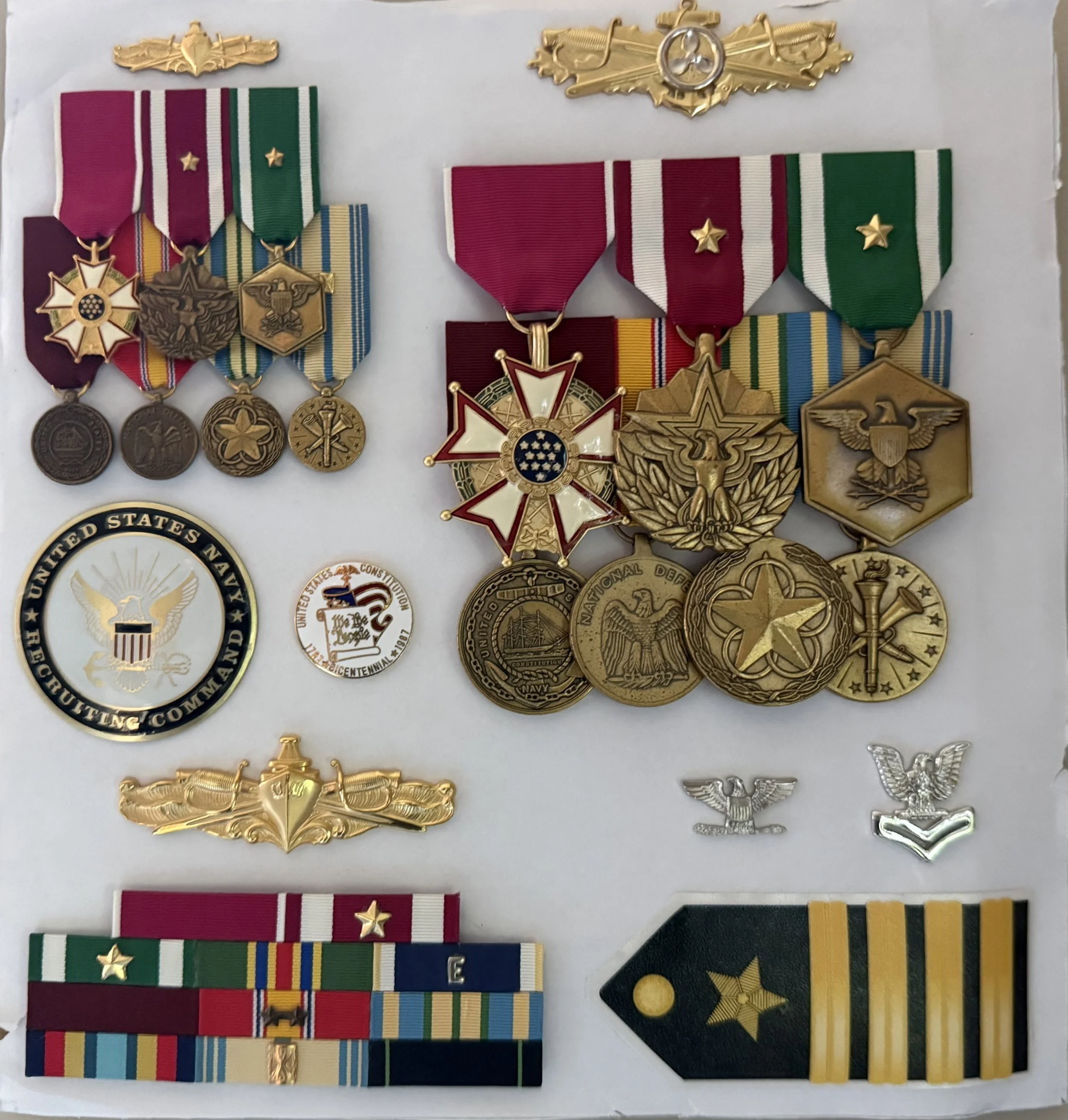 Display of various military service medals and ribbons on a white background earned by CAPT Curt Hess during his 36-years of uniformed service in the USNavy. Also included: Surface Warfare and Surface Engineering Duty Officer pins