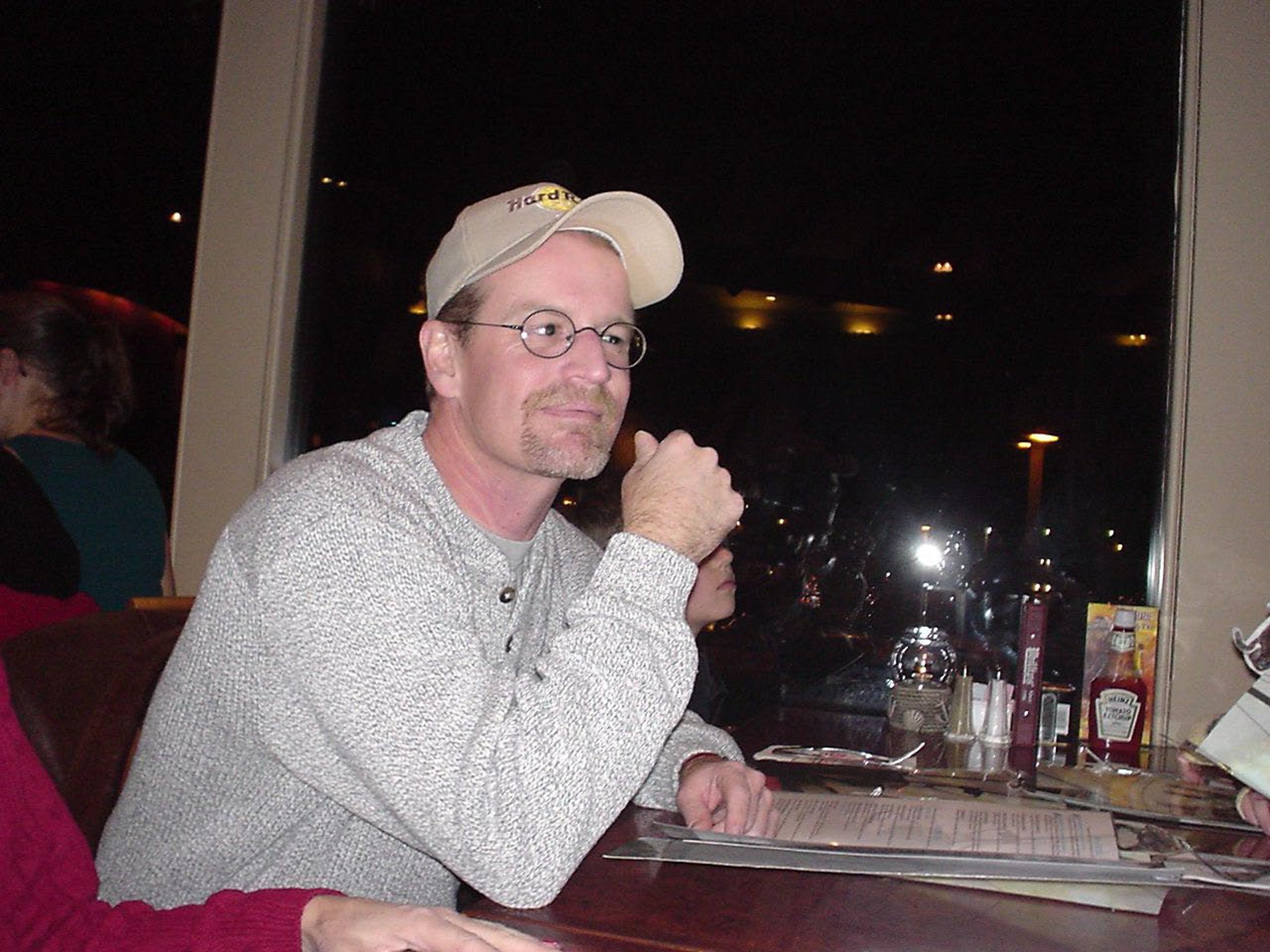 Karl Hess, wearing a beige cap sitting at a restaurant table with drinks, condiments, and a menu, looking to the right at night.