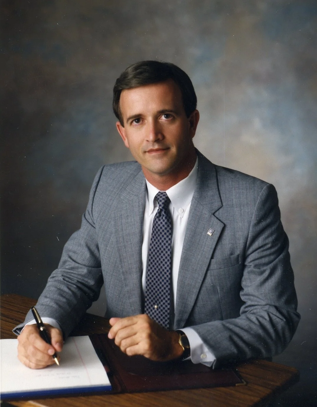 Portrait of businessman Curt Hess in a gray suit, white shirt, and patterned tie, sitting at a wooden desk with a pen and clipboard, against a blurred background.