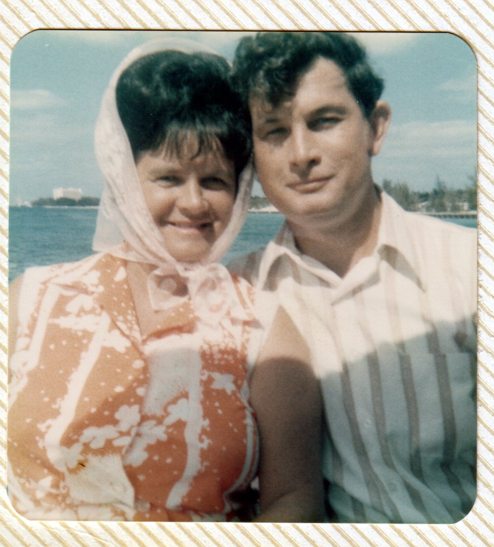 A vintage photo of a smiling woman wearing a pink patterned dress with a matching scarf, standing next to a man in a light-colored striped shirt, outdoors near a body of water with buildings and trees in the background.