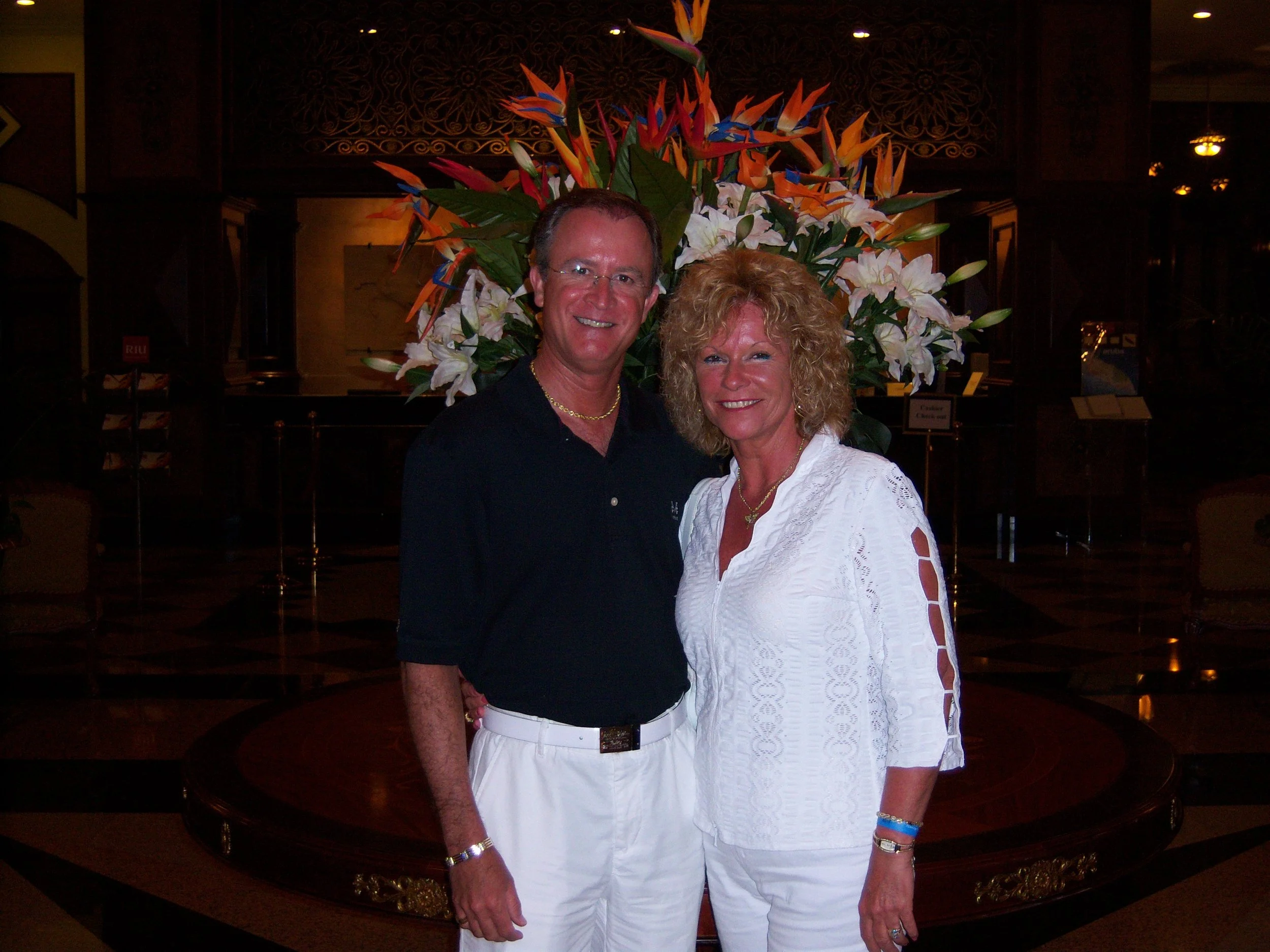Stan Hess and his wife Nancy pose in front of a large floral arrangement featuring orange, white, and blue flowers inside a dimly lit indoor setting.