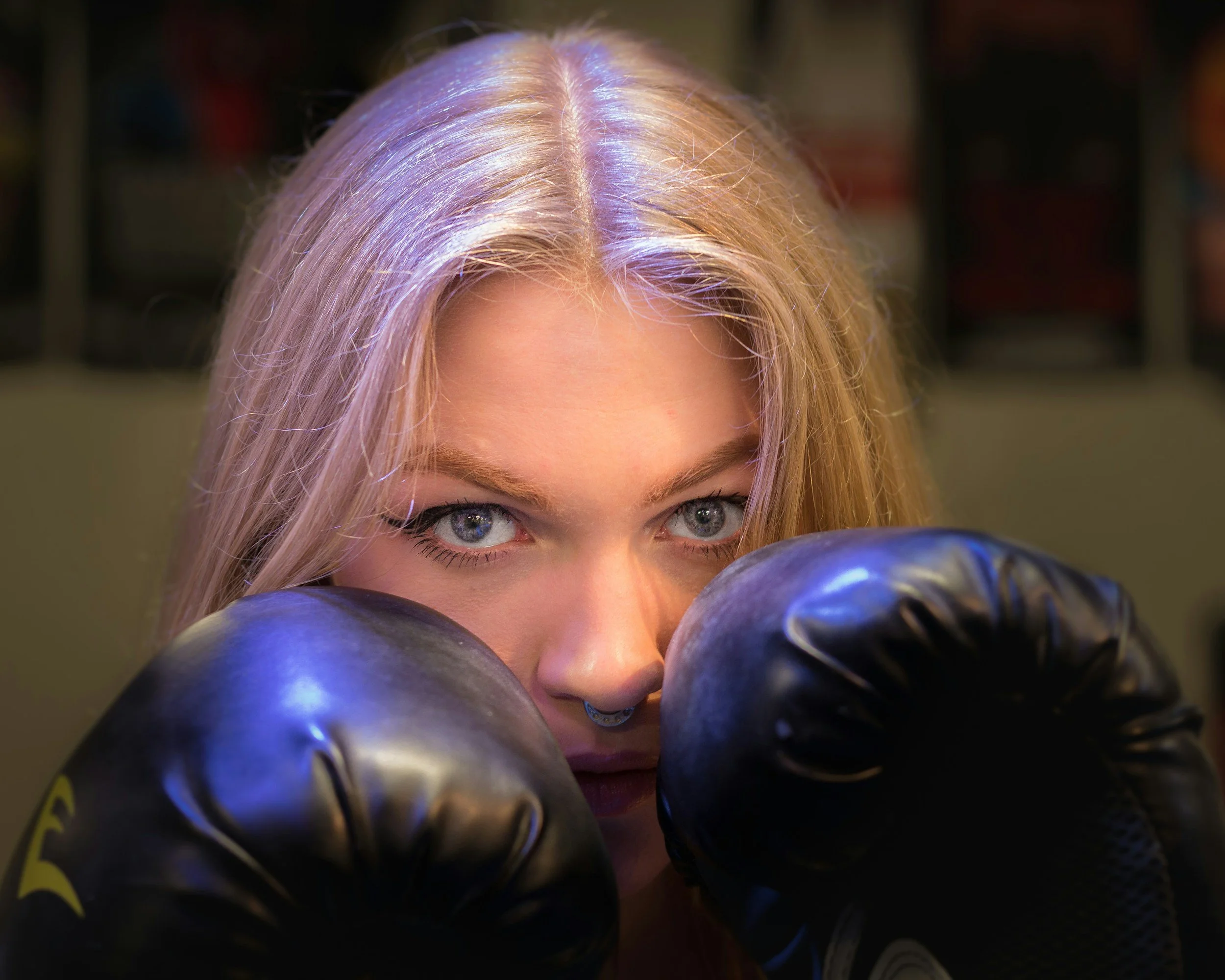 Close-up of a woman's face with blonde hair and blue eyes, surrounded by several shiny, metallic boxing gloves.
