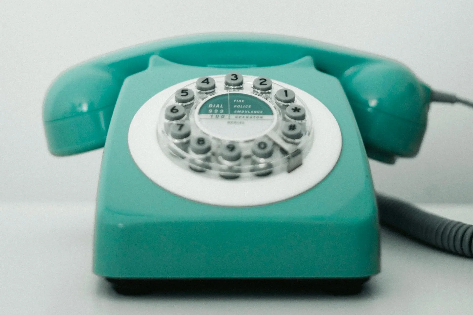 A turquoise rotary telephone with a rounded dial and a coiled cord, sitting on a white surface.