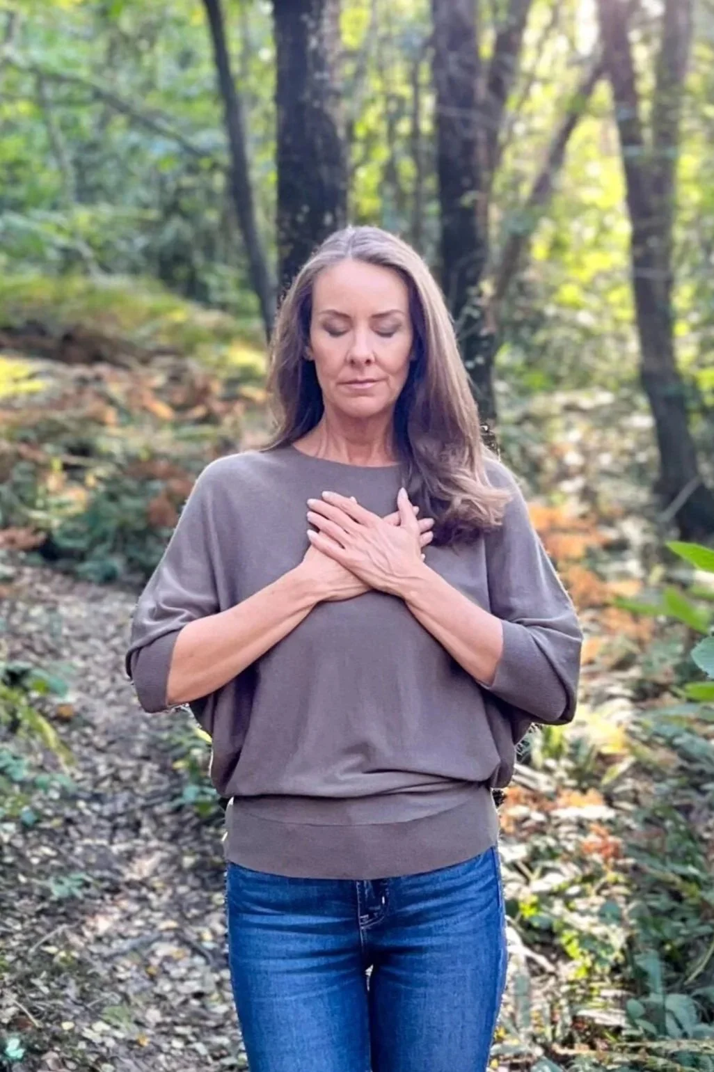 A woman with long brown hair stands in a forest, with eyes closed and hands on her chest, appearing to meditate or reflect peacefully.