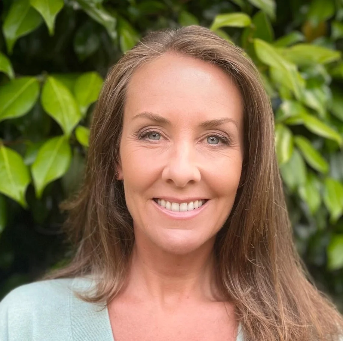 A woman with long light brown hair and blue eyes smiling outdoors with green leafy plants in the background.