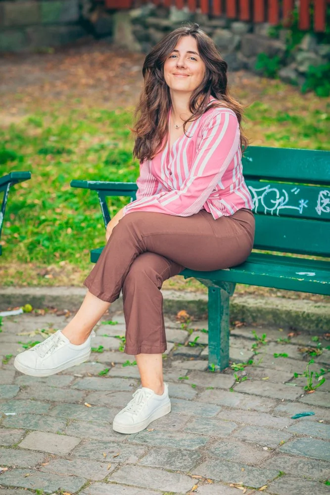 En kvinna som sitter på en parkbänk utomhus, ler och har långt, mörkt hår, bär rosa och vita randiga blusar, bruna byxor och vita sneakers. Bakgrund med grönt gräs och träd.