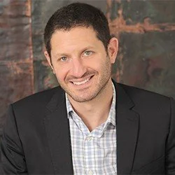 A man with short dark hair and a beard, wearing a black blazer over a light checkered shirt, smiling against a brick wall background.