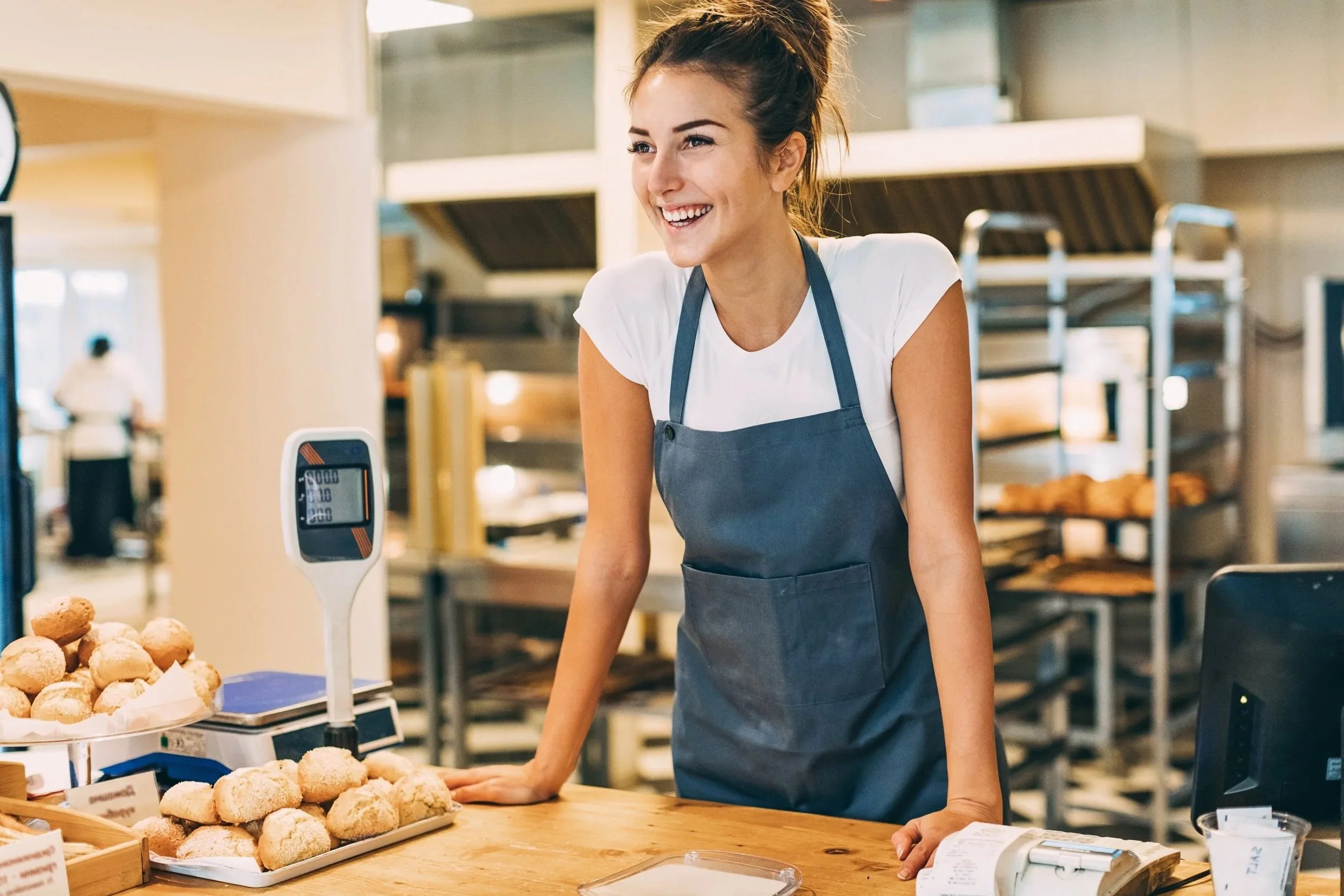 A young woman with a happy expression, wearing a white t-shirt and a gray apron, working at a bakery counter with baked goods and a digital scale, in a bakery or cafe setting.