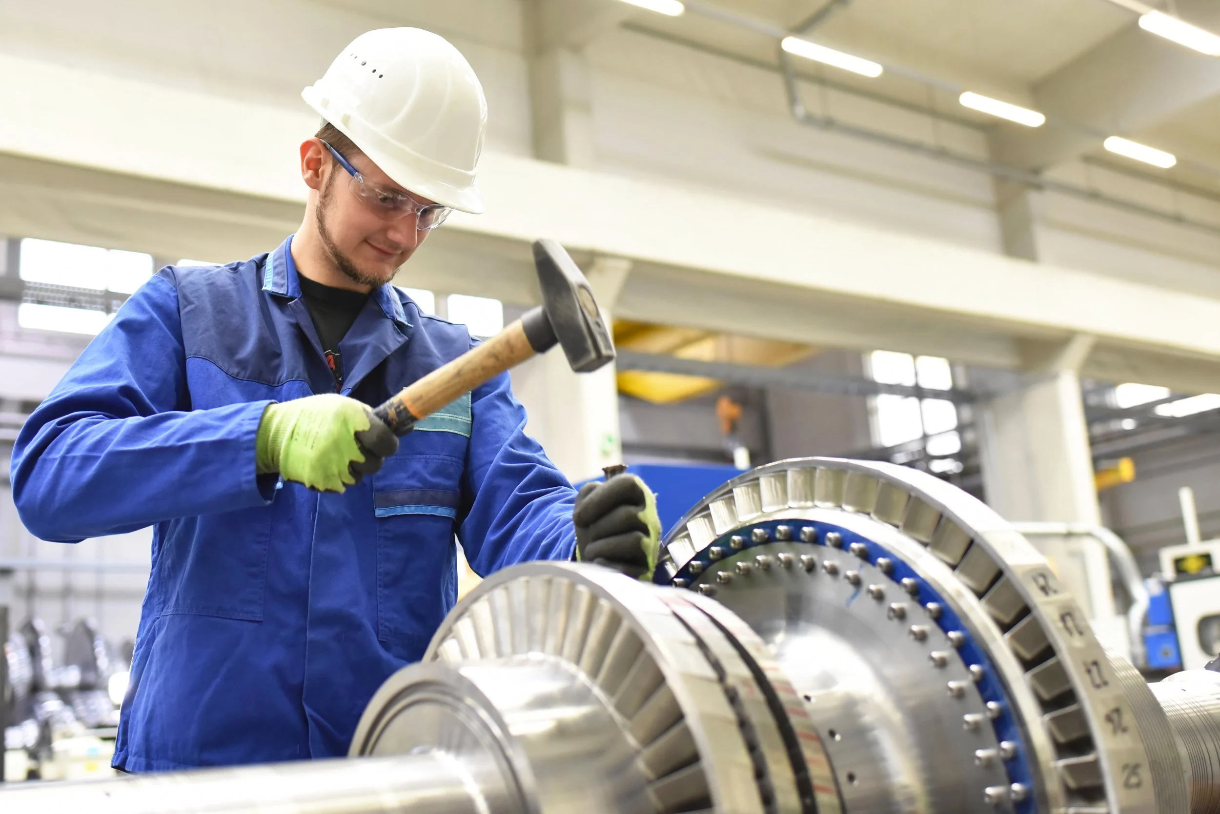 A worker in a blue uniform, safety glasses, and a white hard hat working with machinery in an industrial setting.