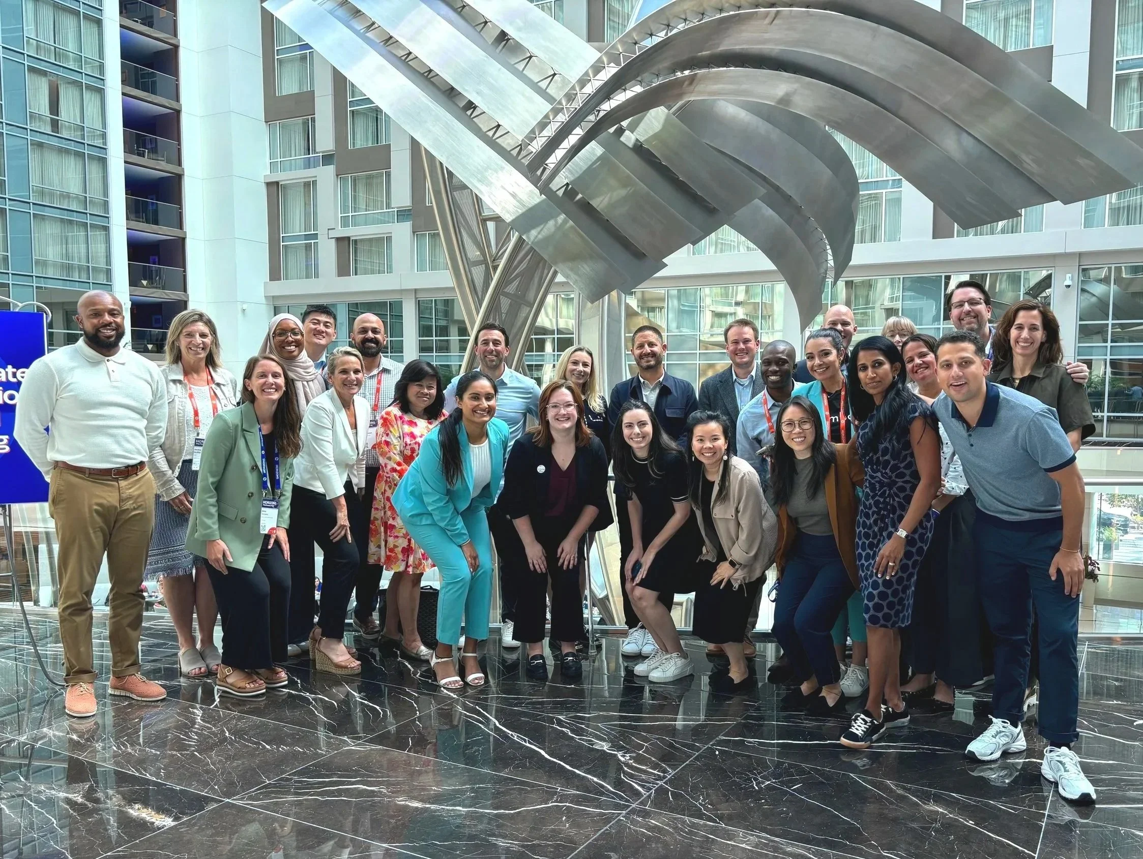 Group of diverse people gathered together in a modern building atrium with large metal sculpture in the background, smiling for the photo.