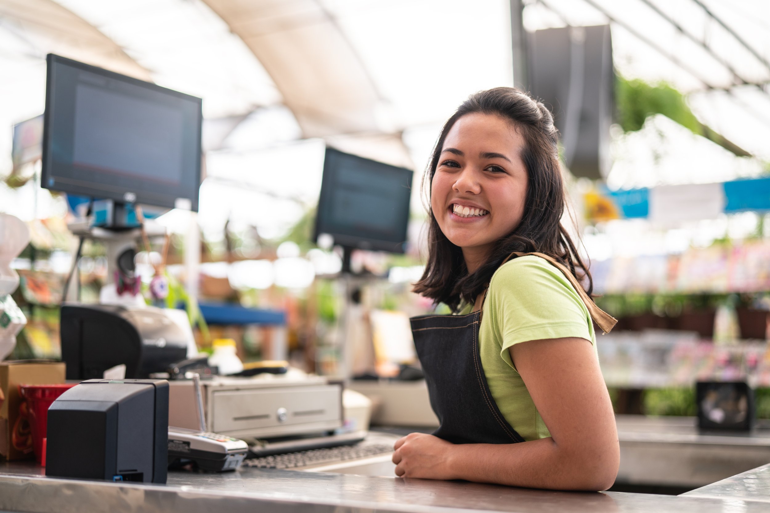 A young woman with dark hair wearing a green shirt and black apron smiling while working at a checkout counter in a bright store with computers and other equipment around her.