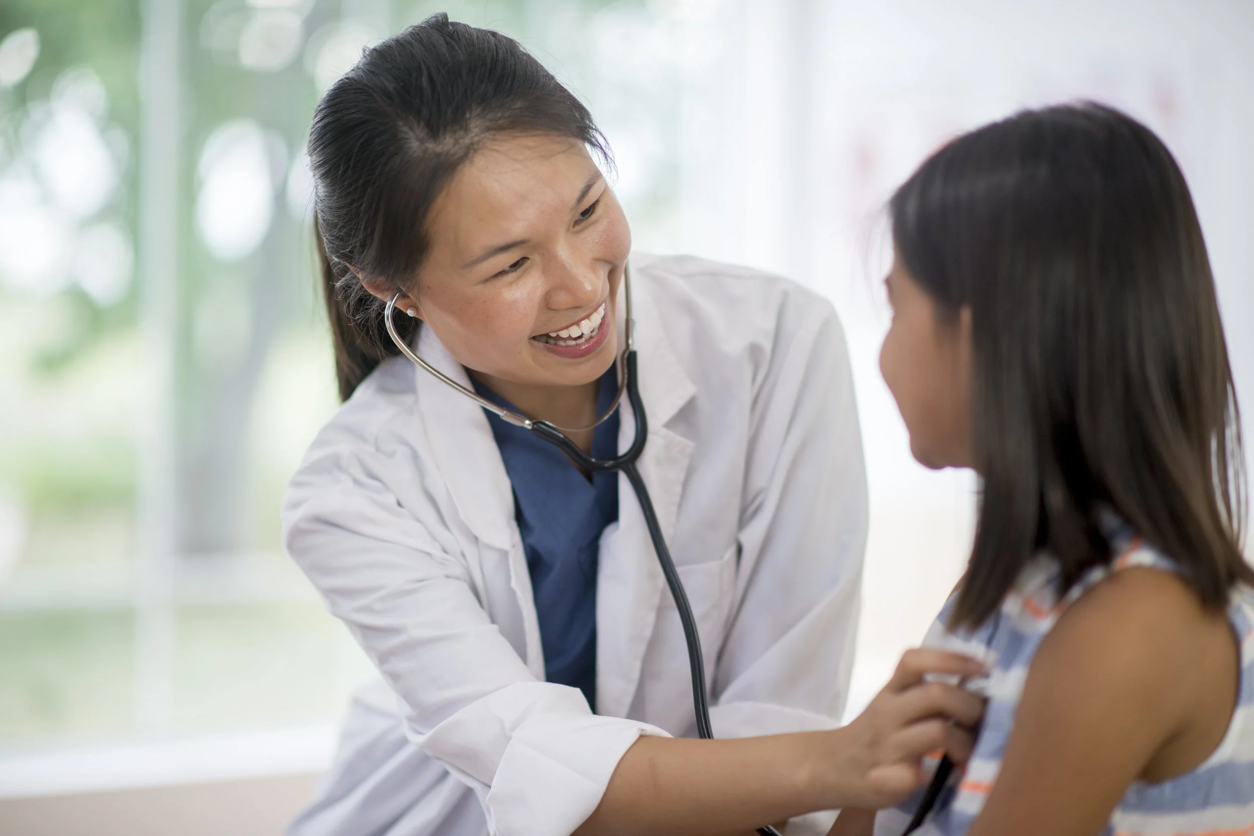 A female healthcare professional in a white coat and stethoscope gently examining a young girl, who appears to be a patient, in a bright, medical setting.