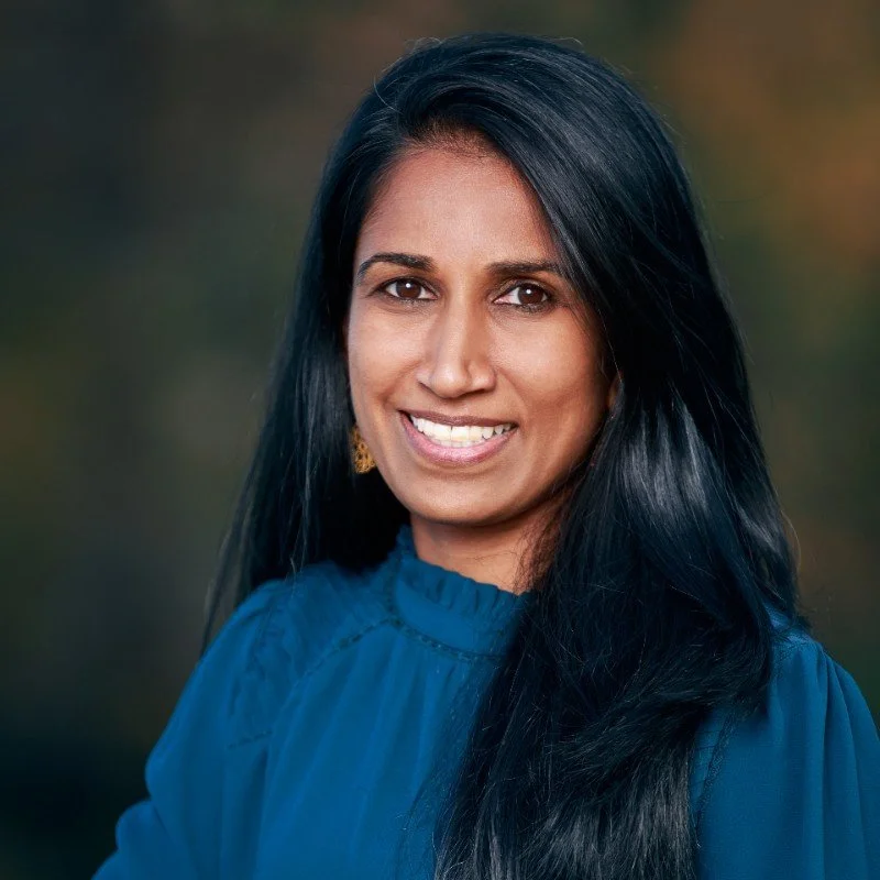 A portrait of a woman smiling outdoors with long black hair, wearing a blue top and gold earrings.