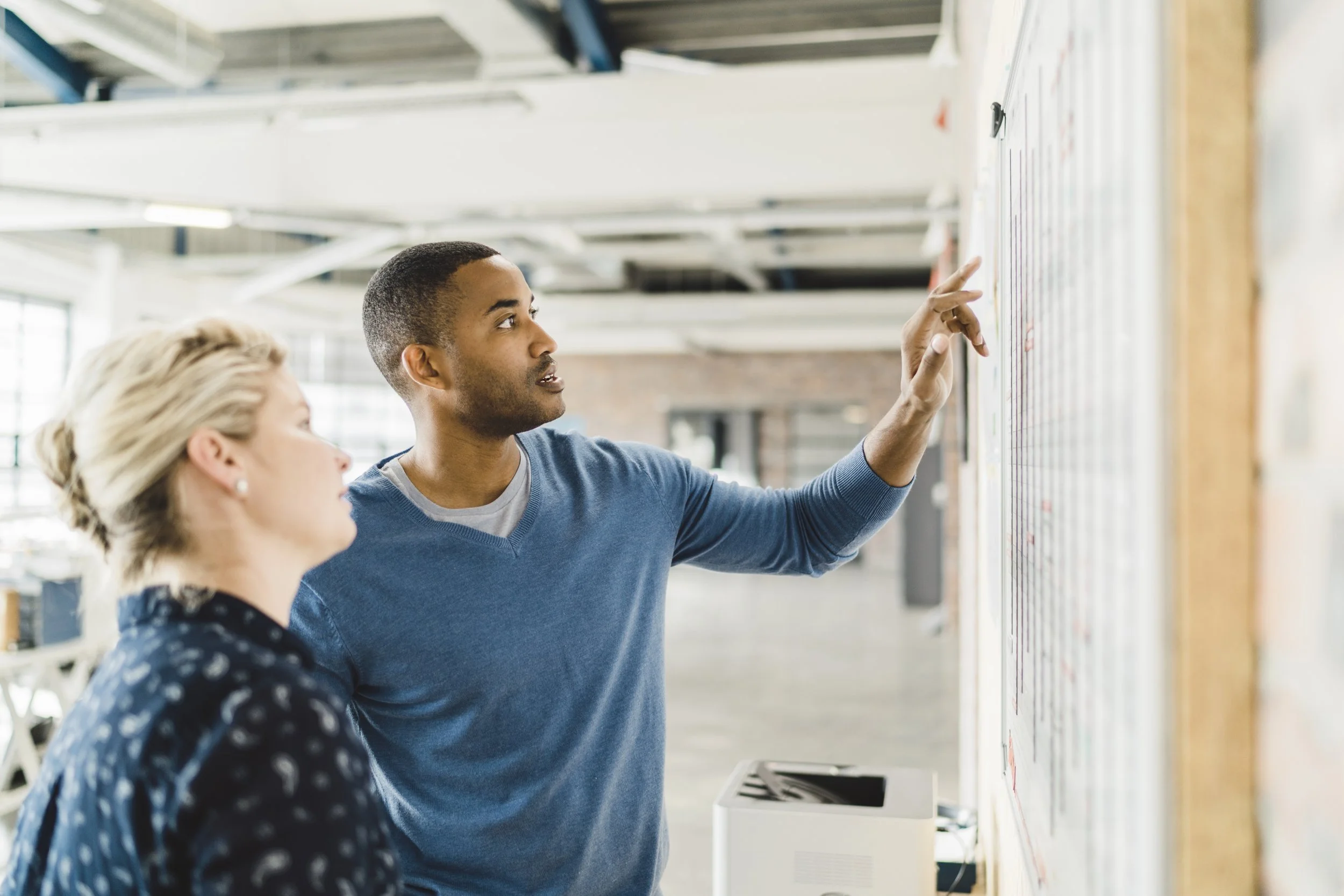A man and a woman are standing in front of a whiteboard with colorful sticky notes. The man is pointing at the whiteboard and appears to be explaining something, while the woman looks on attentively. The setting looks like a modern office or collaborative workspace.