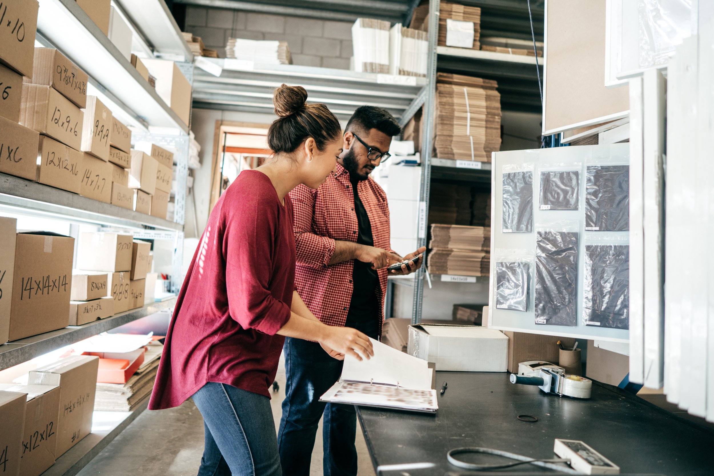 Two people in a warehouse looking at a tablet with shelves of boxes and materials around them.