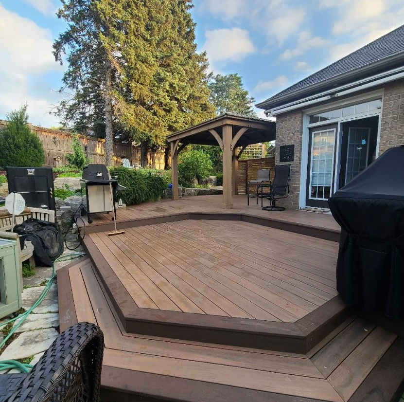 Backyard wooden deck with a rocking chair, barbecue grills, a shaded sitting area, and surrounding greenery including trees and bushes.