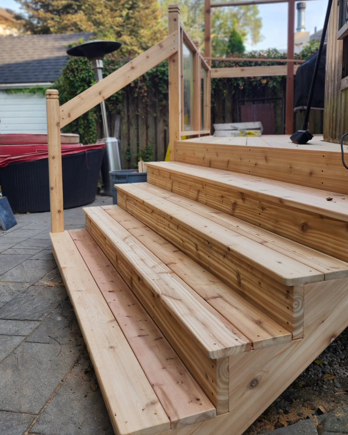 Wooden stairs under construction in a backyard, with a railing partially installed. There is an outdoor heater, a hot tub, and garden tools nearby.