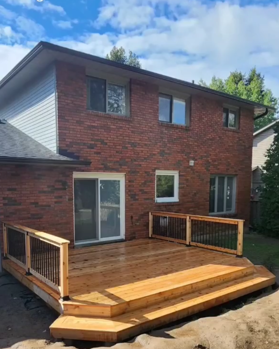 Newly built wooden deck attached to a brick house with sliding glass doors and multiple windows, surrounded by a backyard with trees.